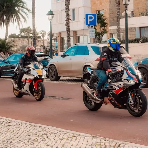 Twee motorrijders rijden achter elkaar door druk stadsverkeer langs geparkeerde auto's, vergelijkbaar met de route tijdens het AVD examen